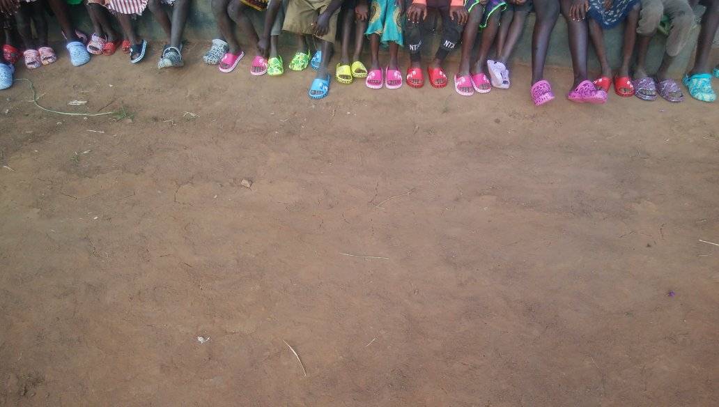 A long row of children's legs and feet, every child in bright new sandals, on red earth